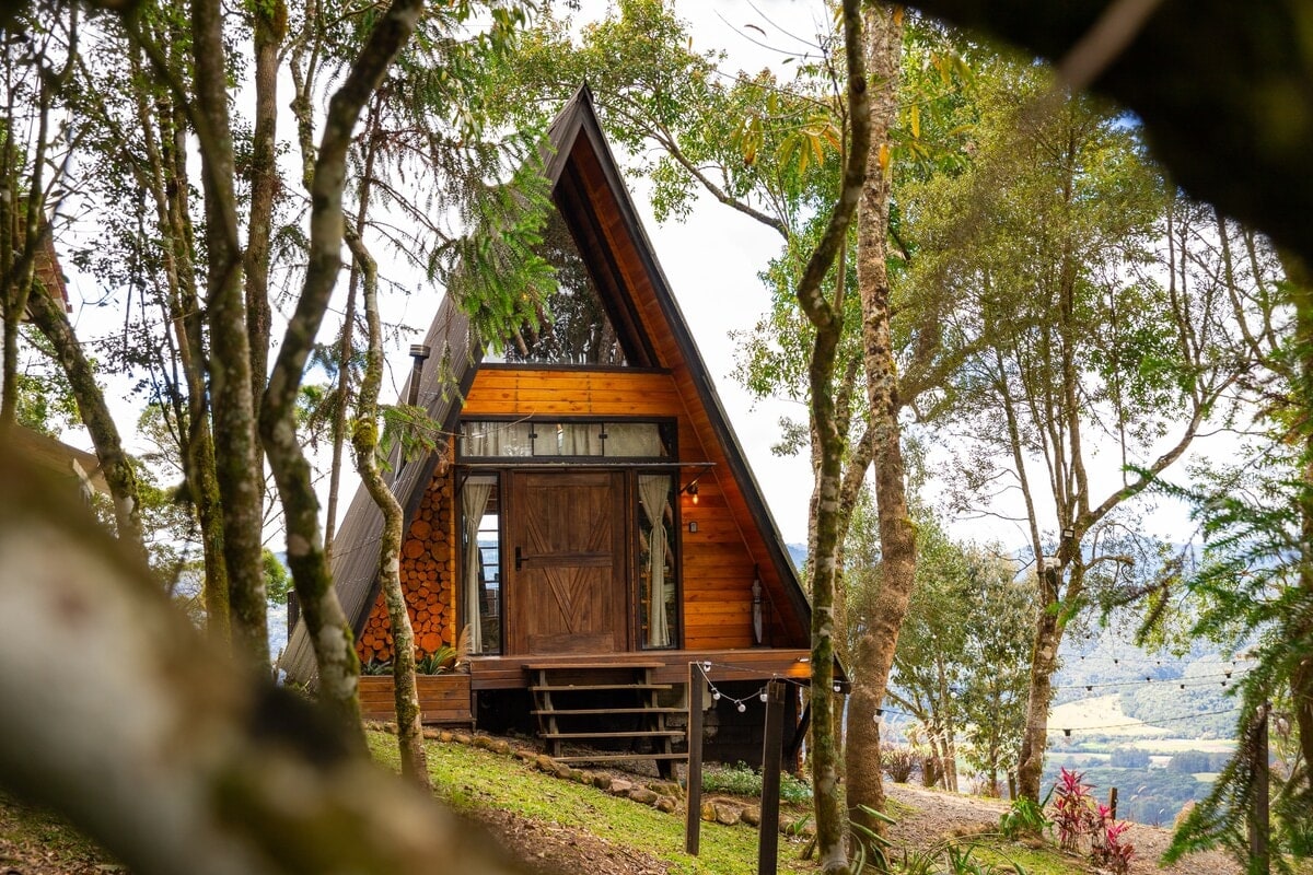 Um chalé A-frame de madeira entre árvores, com vista para o vale, como foi anunciado, de dia sob um céu claro.